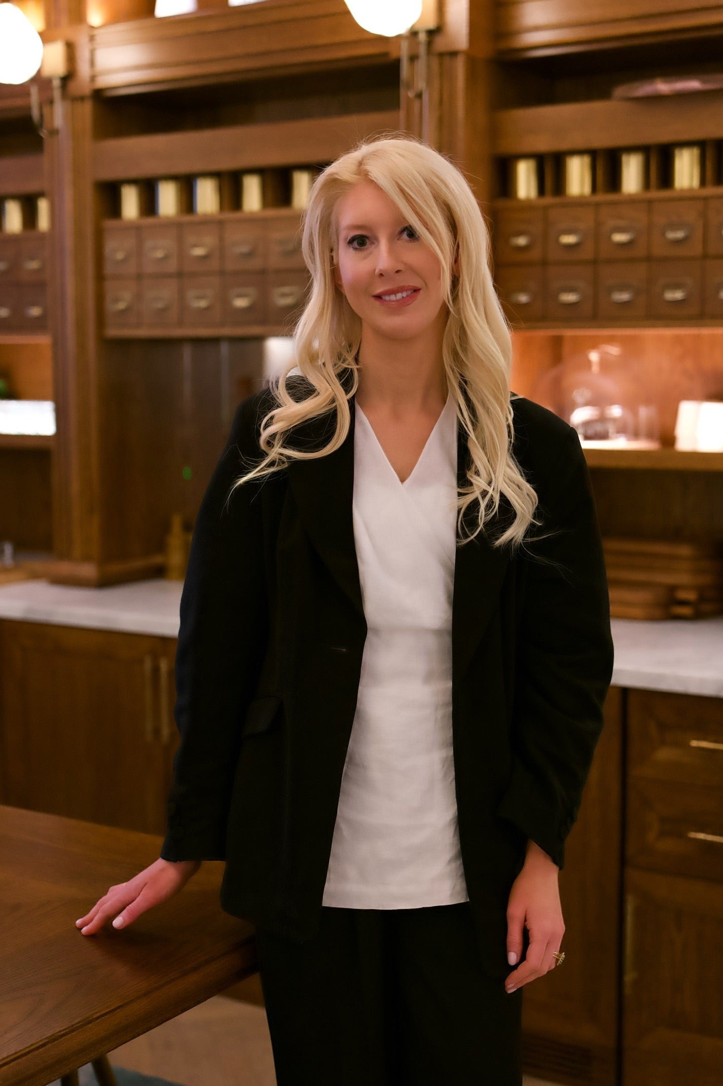Bella von Nesselrode at Six Senses London in a black blazer and white shirt standing in a room with apothcary cabinets.